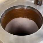 Tandoor oven Curing Paste AS05 – overhead view inside a stainless steel-jacketed tandoor oven showing the clay interior walls fully coated with a layer of dark brown curing paste, with the AS05 Curing Paste jar partially visible in the top right corner, prior to the first seasoning burn