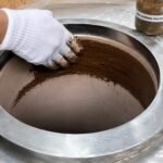 Tandoor oven Curing Paste AS05 – gloved hand rubbing dark brown curing paste onto the inner clay walls of a stainless steel-jacketed tandoor oven, with the AS05 Curing Paste jar visible in the top right corner, demonstrating the hand-application method used during the tandoor seasoning process