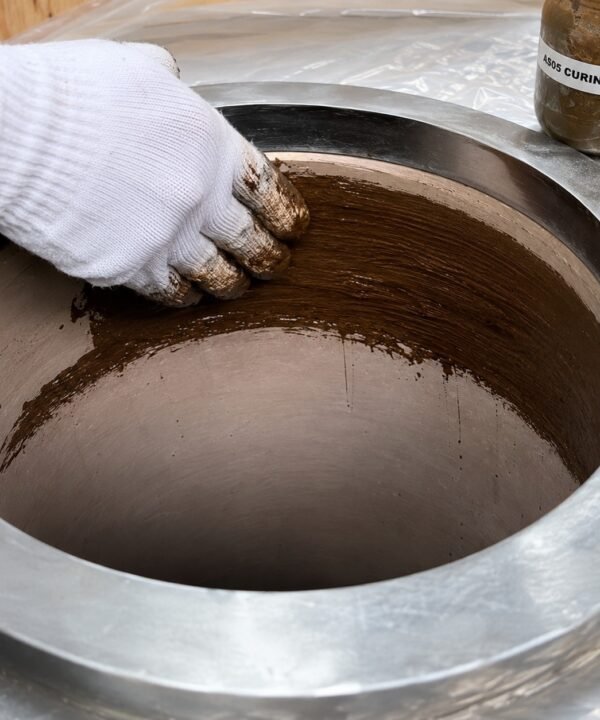 Tandoor oven Curing Paste AS05 – gloved hand rubbing dark brown curing paste onto the inner clay walls of a stainless steel-jacketed tandoor oven, with the AS05 Curing Paste jar visible in the top right corner, demonstrating the hand-application method used during the tandoor seasoning process