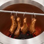 Four Tandoor Meat Hanging Hook AS17 units suspending marinated lamb chops from a horizontal rod inside a stainless steel tandoor oven over glowing red embers