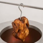 Tandoor Meat Hanging Hook AS17 holding a whole tandoori roasted chicken by its cavity above the opening of a stainless steel tandoor oven
