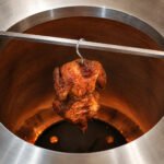 Tandoor Meat Hanging Hook AS17 suspending a spiced whole chicken deep inside a stainless steel tandoor oven with orange flames glowing at the bottom