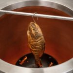 Tandoor Meat Hanging Hook AS17 suspending a whole roasted fish vertically inside a glowing clay tandoor oven with live embers visible at the base