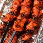 Close-up of multiple black flat Tandoor Skewers loaded with charred red chicken tikka pieces resting on a foil-lined tray