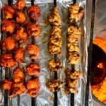 Four black flat Tandoor Skewers carrying red and white marinated chicken tikka pieces resting on a foil tray beside a glowing tandoor oven