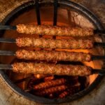 Four flat Tandoor Skewers loaded with grilled seekh kebabs resting horizontally across the top of a tandoor oven opening with glowing coals and char visible beneath