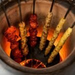 Multiple Tandoor Skewers loaded with assorted kebabs including red chicken tikka, seekh kebab, and yellow paneer standing vertically inside a glowing clay tandoor oven with bright orange embers at the base