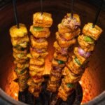 Four flat Tandoor Skewers loaded with spiced paneer tikka cubes and mixed vegetables including red onion and green pepper cooking inside a glowing clay tandoor oven