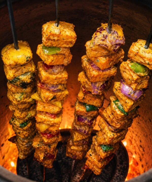Four flat Tandoor Skewers loaded with spiced paneer tikka cubes and mixed vegetables including red onion and green pepper cooking inside a glowing clay tandoor oven