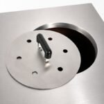 Stainless Steel Tandoor Oven Lid AS11 – angled close-up view of the stainless steel lid being slid partially open on a tandoor oven top surface, showing the black heat-resistant bar handle, ventilation holes, and the dark oven mouth opening revealed beneath the lid edge