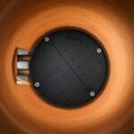 Tandoor Baffle Plate AS02 – overhead view of baffle plate installed inside a tandoor clay pot, showing the round black steel plate with Morni brand name etched into surface, perforated holes, and centre divider rod resting against warm orange clay walls with burner pipes visible to the left