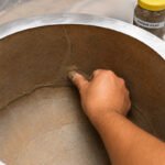 Tandoor Clay Pot Repair Kit AS08 – hand using thumb to press and smooth repair clay into a crack running along the inner clay wall of a stainless steel-jacketed tandoor oven, with the Repair Clay jar visible in the top right corner, demonstrating the crack-filling application process