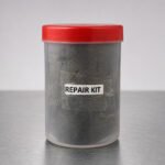 Tandoor Clay Pot Repair Kit AS08 – front view of clear cylindrical plastic container with red lid containing dark grey repair clay compound, showing REPAIR KIT label with label edges peeling, placed on a brushed stainless steel surface in a minimal studio setting