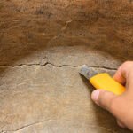Tandoor Clay Pot Repair Kit AS08 – close-up of a hand using a yellow-handled scraping tool to clean out and open up a crack on the inner clay wall of a tandoor oven, showing the surface preparation step before applying repair clay