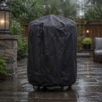Tandoor Cover AS14 – black all-weather tandoor cover fully fitted and drawstring-secured over a tandoor oven on casters, standing on a wet stone patio during heavy rainfall, with water droplets beading on the waterproof cover surface, a stone lantern pillar to the left, outdoor seating and warm patio lighting visible in the background, demonstrating the cover's rain protection in real outdoor conditions