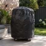Tandoor Cover AS14 – black all-weather tandoor cover fully fitted over a tandoor oven on casters, placed on a patio stone surface in a landscaped backyard setting with green plants, wood fence, and outdoor seating visible, showing the cover protecting the tandoor from outdoor elements in warm weather
