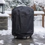Tandoor Cover AS14 – black all-weather tandoor cover fully fitted and drawstring-secured over a tandoor oven on casters, standing on a snow-covered stone patio in winter conditions, with snow-laden trees, iron railings, and a stone lantern visible in the background, demonstrating the cover's all-season outdoor weather protection