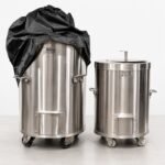 Tandoor Cover AS14 – studio shot showing a black tandoor cover partially draped over a large stainless steel Morni tandoor oven on the left, beside a smaller uncovered stainless steel cylindrical tandoor oven on the right, illustrating the cover's sizing and fit across different tandoor oven sizes