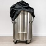 Tandoor Cover AS14 – front view of a black tandoor cover being placed over the top of a stainless steel Morni cylindrical tandoor oven on casters, showing the cover bunched at the top mid-placement with the Morni branding, side handles, and ash drawer on the oven body still visible below