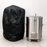 Tandoor Cover AS14 – studio shot on white background showing the black tandoor cover standing upright on the left next to an uncovered stainless steel cylindrical Morni tandoor oven on casters on the right, demonstrating the cover's size relative to the oven it is designed to protect