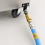 Tandoor Gas Hose AS01 – flexible blue gas hose installed on the base of a stainless steel tandoor oven on casters, with brass quick disconnect coupling engaged at the appliance gas inlet connection point
