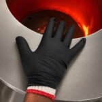 Tandoor Heat Resistant Gloves AS15 – close-up of a gloved hand resting on the stainless steel rim of a tandoor oven opening, with intense orange-red glowing clay walls and visible flame inside the tandoor, showing the black nitrile outer glove and white cotton inner liner cuff at the wrist providing dual-layer protection at high heat