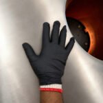 Tandoor Heat Resistant Gloves AS15 – gloved hand placed flat against the brushed stainless steel exterior of a tandoor oven beside the glowing oven opening, showing the full black nitrile outer glove with white and red cotton inner liner cuff visible at the wrist, demonstrating dexterity and fit during tandoor handling