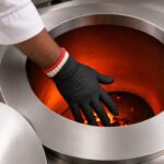 Tandoor Heat Resistant Gloves AS15 – overhead view of a chef's hand wearing the double-layer glove system — black nitrile outer glove over white cotton inner liner with red trim cuff — reaching into a glowing orange-red tandoor oven interior with live flame and charcoal visible at the base, showing the gloves in active high-heat use