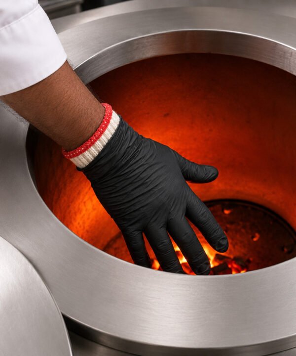 Tandoor Heat Resistant Gloves AS15 – overhead view of a chef's hand wearing the double-layer glove system — black nitrile outer glove over white cotton inner liner with red trim cuff — reaching into a glowing orange-red tandoor oven interior with live flame and charcoal visible at the base, showing the gloves in active high-heat use