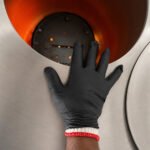 Tandoor Heat Resistant Gloves AS15 – gloved hand wearing the double-layer system reaching into the opening of a stainless steel-jacketed commercial tandoor oven, with the glowing orange clay interior and lit Morni baffle plate visible inside, showing the gloves protecting the hand at the oven mouth during tandoor operation