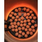 Tandoor Oven Clay Balls AS07 – overhead angled view of clay balls arranged across a Morni-branded tandoor baffle plate installed inside a glowing orange tandoor oven, with the burner pipes visible to the lower left and heat radiating from the terracotta clay walls