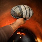 Tandoor Oven Naan Gaddi AS09 – hand gripping a dark green and black plaid naan gaddi cushion inside a glowing orange-hot tandoor oven, with the Morni-branded baffle plate visible below and radiant heat from the clay walls surrounding the gaddi