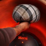 Tandoor Oven Naan Gaddi AS09 – hand holding a dark plaid woven naan gaddi inside a fully heated tandoor oven, with the glowing red-orange clay walls and the Morni baffle plate visible below, showing the gaddi in its working environment at high temperature