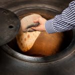 Tandoor Oven Naan Gaddi AS09 – hand using a worn striped naan gaddi to press and slap a raw naan dough portion onto the inner clay wall of a traditional black iron-clad tandoor oven, with the oven lid partially open and the clay interior glowing orange from heat