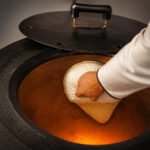 Tandoor Oven Naan Gaddi AS09 – chef's hand using a plain white cotton naan gaddi to press and flatten raw naan dough onto the hot inner clay wall of a tandoor oven, with the black iron lid propped open and the clay interior glowing warm amber from the burner