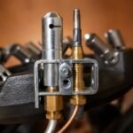 Tandoor Oven Thermocouple AS12 – close-up of the thermocouple probe and pilot assembly bracket mounted on a cast iron tandoor burner, showing the stainless steel pilot nozzle tube, brass gas fittings, thermocouple probe tip with copper capillary lead, igniter electrode, and steel mounting bracket, with burner nozzle tips visible in the blurred background