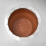 Tandoor Skewers Ring AS10 – overhead view of the stainless steel skewers ring installed around the opening of a fresh terracotta clay-lined tandoor pot, showing the ring resting on the brushed stainless steel oven top with all skewer notch slots visible and the warm orange clay pot interior below