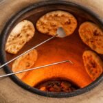 Tandoori Naan Rod Pair AS03 – both rods in use inside a glowing tandoor oven; the spatula-tip rod is lifting a freshly baked naan off the clay wall while the hook-tip rod is positioned to peel another, with four naan breads visible stuck to the terracotta interior walls above a bright orange fire