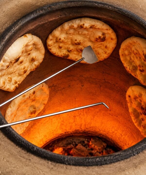 Tandoori Naan Rod Pair AS03 – both rods in use inside a glowing tandoor oven; the spatula-tip rod is lifting a freshly baked naan off the clay wall while the hook-tip rod is positioned to peel another, with four naan breads visible stuck to the terracotta interior walls above a bright orange fire