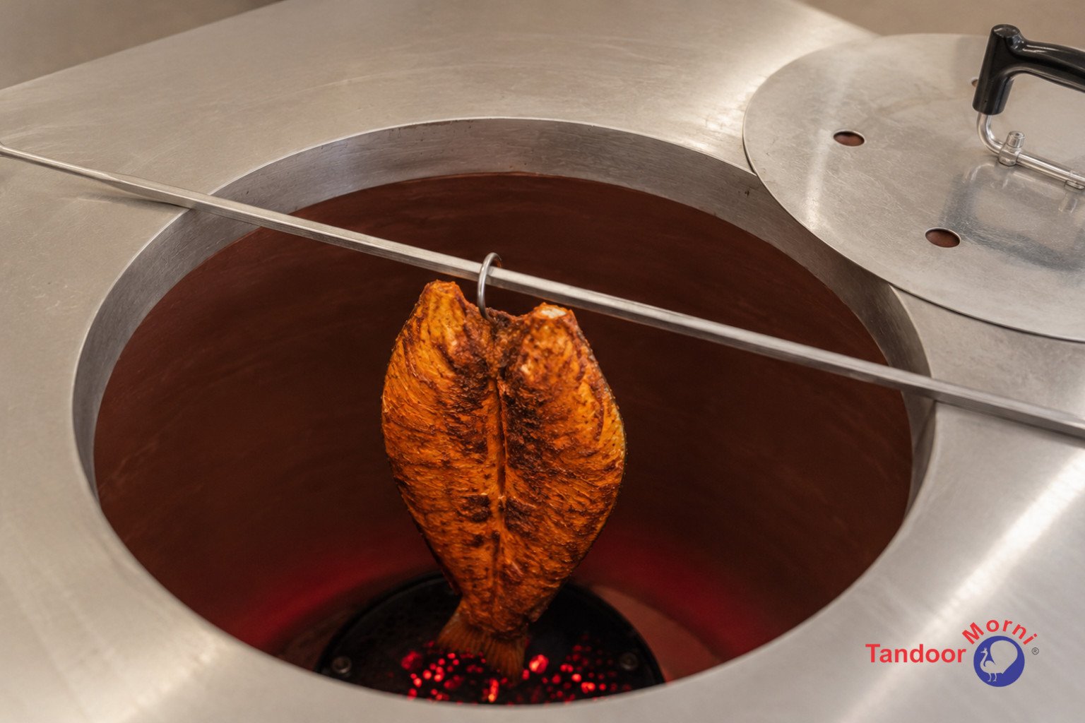 Common mistakes cooking fish in tandoor: a hand lowering a marinated whole fish onto a skewer beside a glowing tandoor oven